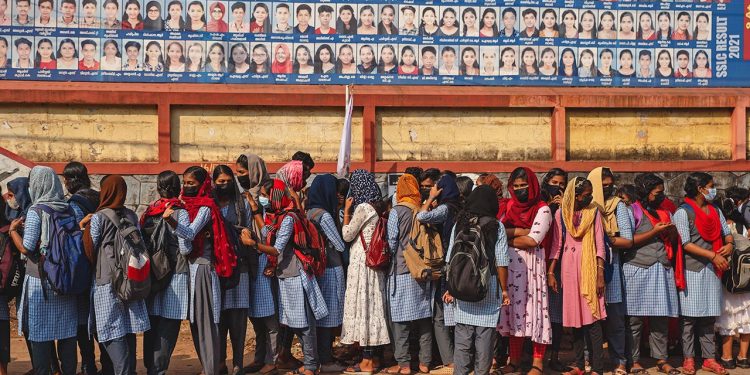 School children await bus transport beneath a billboard with photographs of successful students outside a school in Nooranad, Kerala, India, Friday, March 4, 2022. Slowly but steadily, life in South Asia is returning to normal, and people hope the worst of the COVID-19 pandemic is behind them. (AP Photo/R S Iyer)